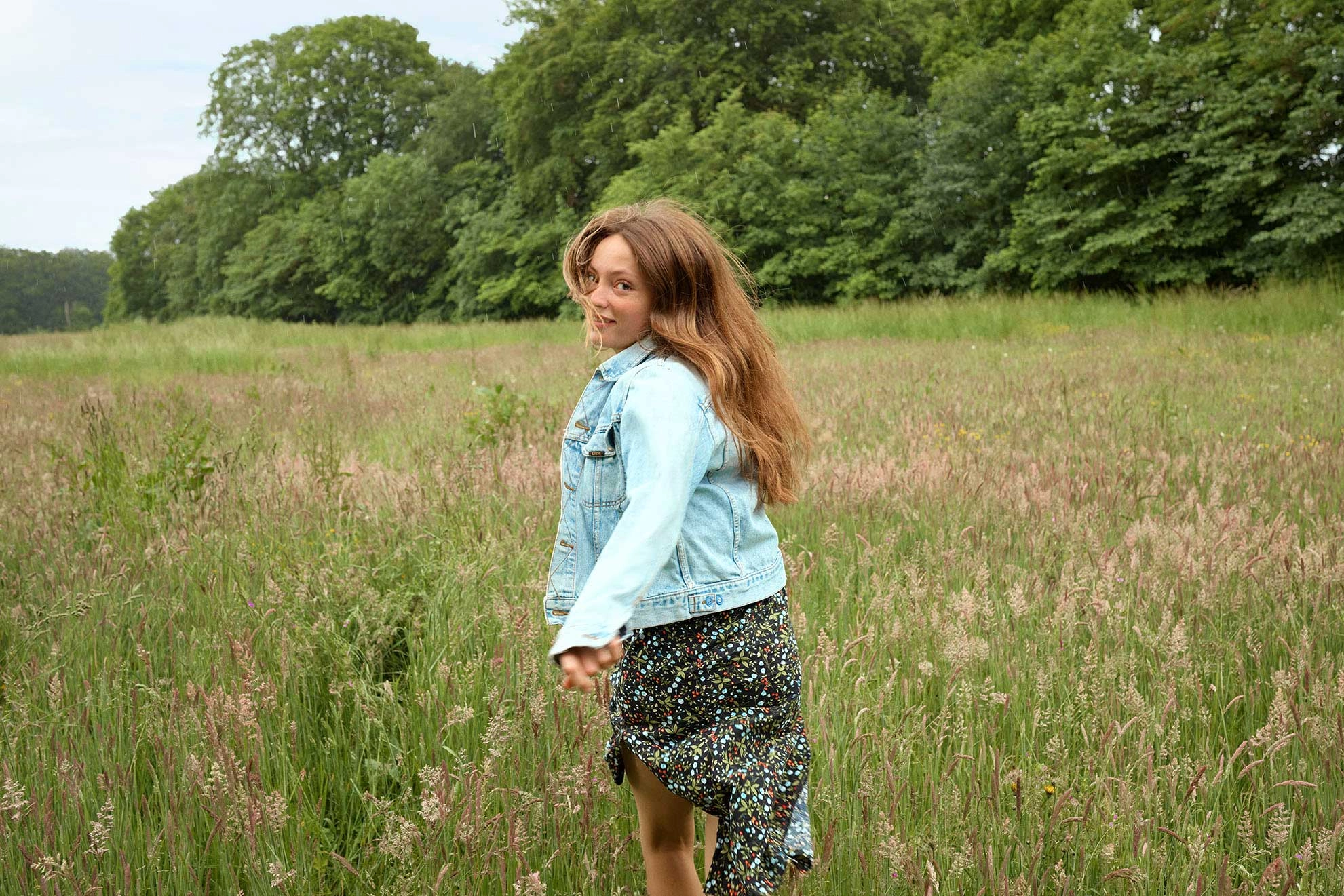 En pige løber i en blomstermark, hun smiler og kigger tilbage mod kameraet