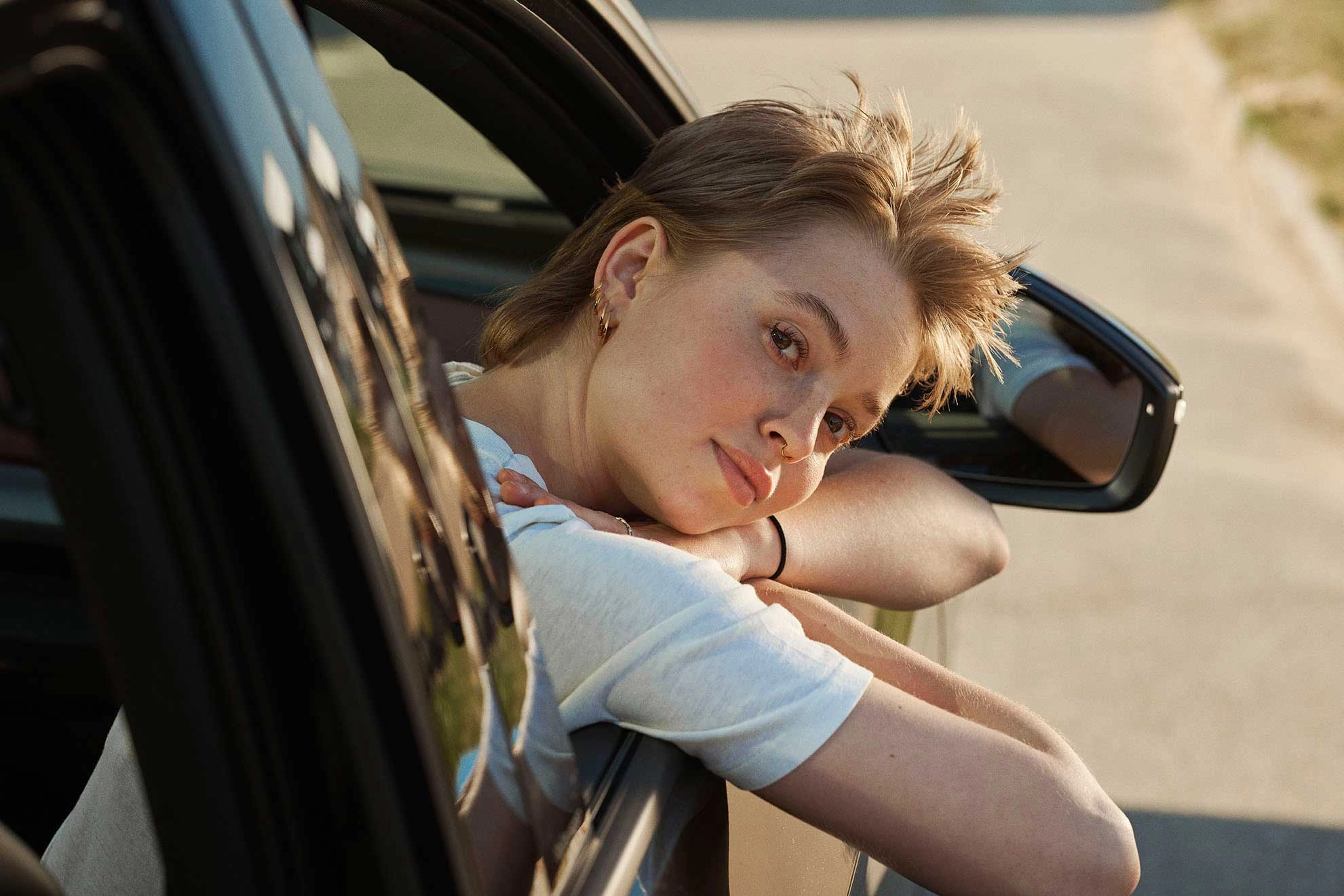 En kvinde sidder i passagersiden af en bil, vinduet er rullet ned og hun sidder med lukkede øjne og nyder solen