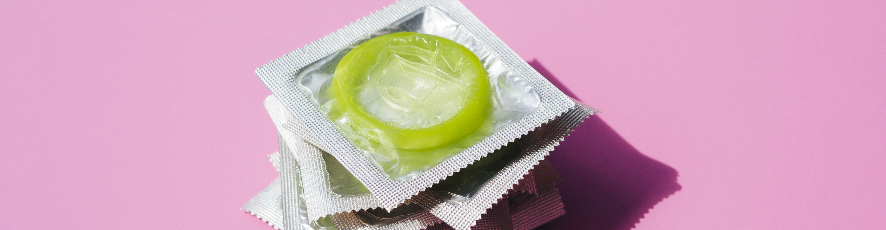 A lime green condom in its foil packaging placed on top of a small stack of individually wrapped condoms, set against a pink background.