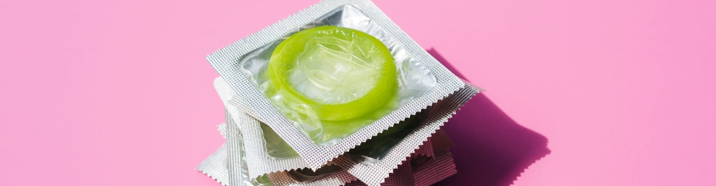 A lime green condom in its foil packaging placed on top of a small stack of individually wrapped condoms, set against a pink background.