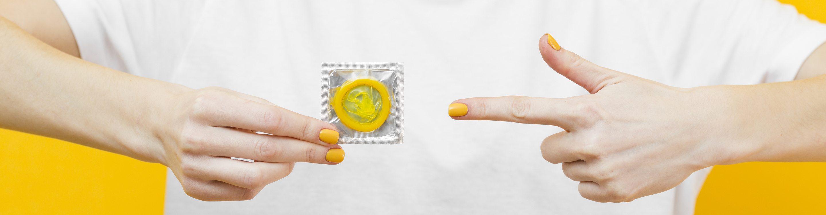 Close-up of two hands against a bright yellow background, one holding a packaged yellow condom and the other pointing toward it.
