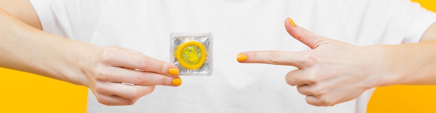 Close-up of two hands against a bright yellow background, one holding a packaged yellow condom and the other pointing toward it.