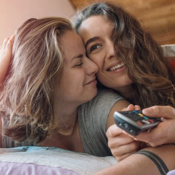 Two girls cuddling and smiling under the covers while holding out a remote control