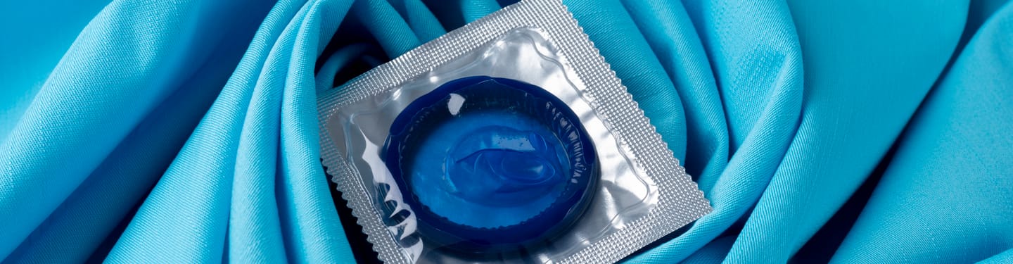 Blue condom in foil packaging placed on soft light blue fabric background, symbolizing sexual health and protection