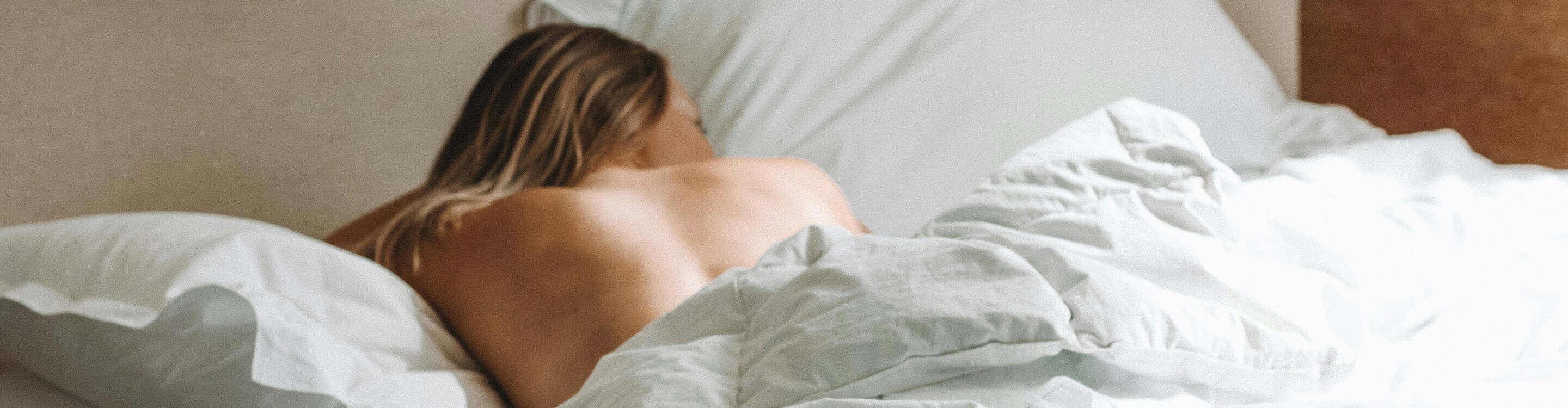 Person lying face down on a bed, partially covered with white bedding in a softly lit room.
