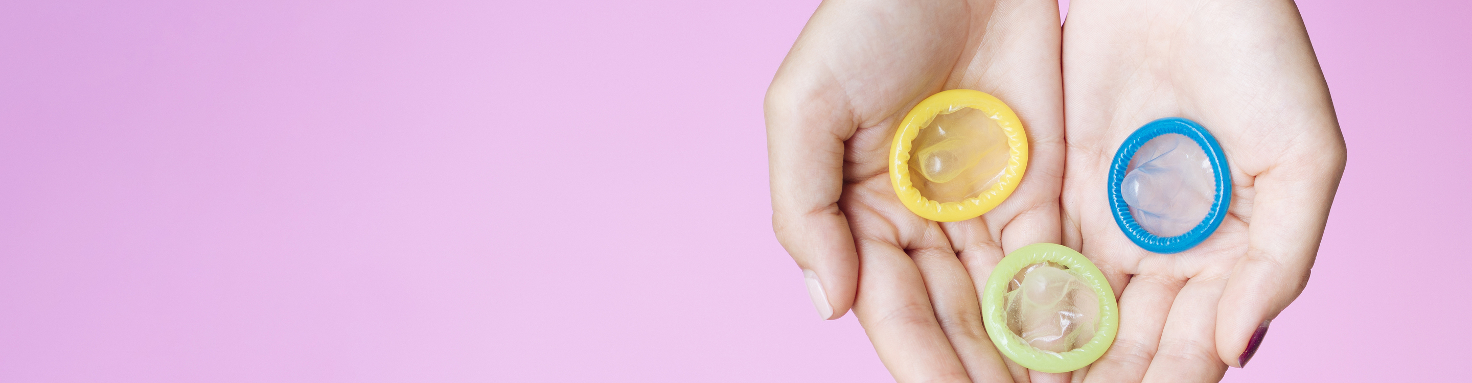Hands holding three different colored condoms (yellow, blue, and green) against a pink background.