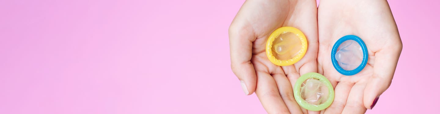 Hands holding three different colored condoms (yellow, blue, and green) against a pink background.