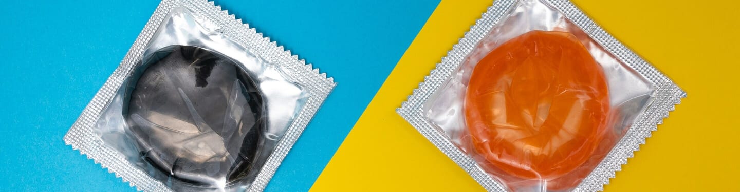 Two individually wrapped condoms in foil packaging, one black and one orange, placed on a blue and yellow background.