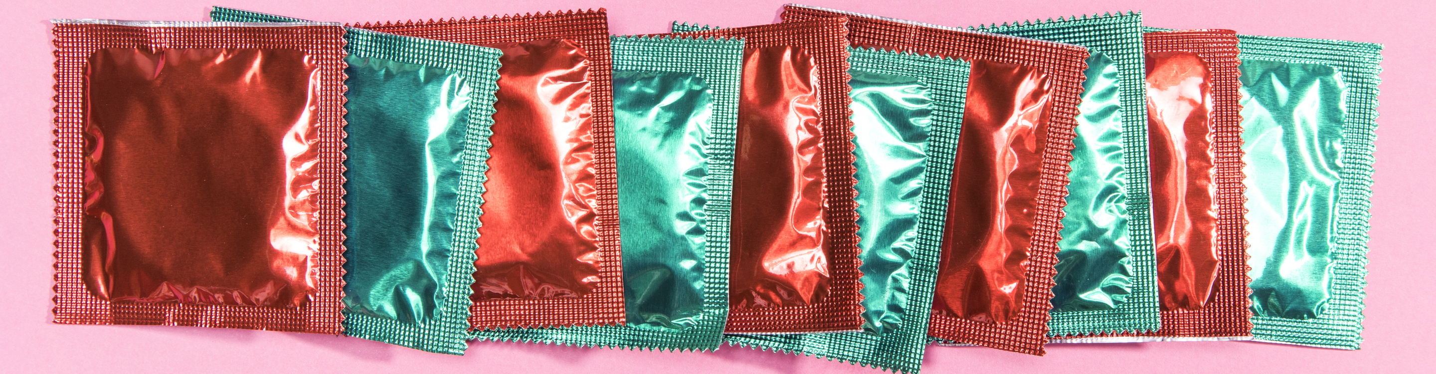 Row of brightly colored foil condom packets in red and green arranged side by side on a pink background