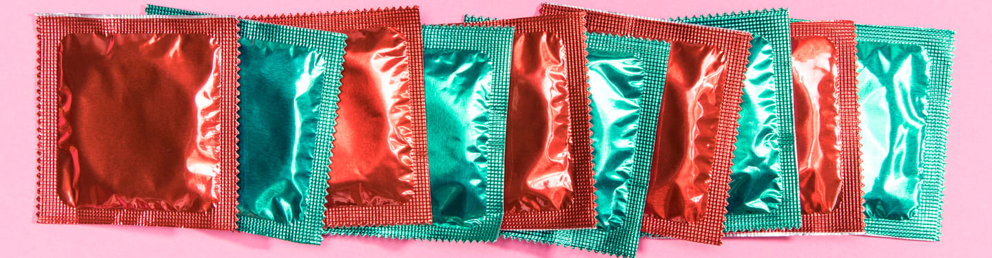 Row of brightly colored foil condom packets in red and green arranged side by side on a pink background