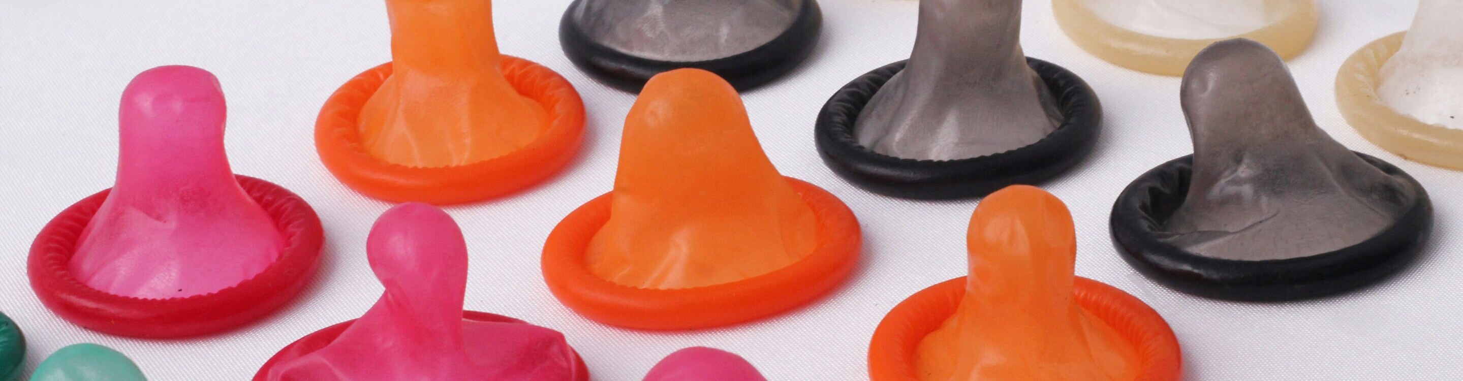 A collection of colorful condoms in various shapes and materials, displayed on a white surface, including options in pink, red, orange, black, and translucent tones.