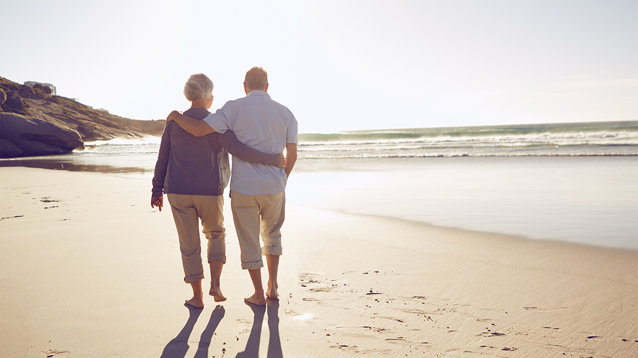 Zwei Personen gehen dicht beieinander am Strand entlang, während sie sich am Rücken und an den Schultern halten. Im Hintergrund sind Meer, Wellen und Felsen zu sehen.
