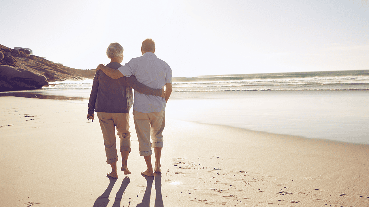 Zwei Personen gehen dicht beieinander am Strand entlang, während sie sich am Rücken und an den Schultern halten. Im Hintergrund sind Meer, Wellen und Felsen zu sehen.
