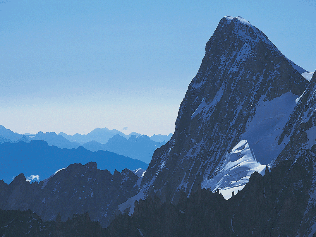 Panoramablick auf eine markante, schneebedeckte Bergspitze Steiler, teils verschneiter Berggipfel mit schattigen Felswänden im Vordergrund und mehreren blauen Bergketten im Hintergrund unter klarem Himmel.