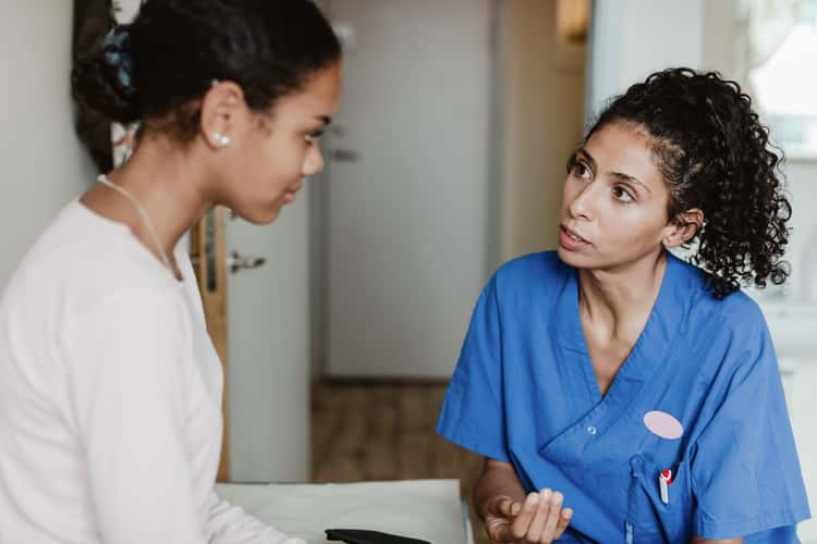 patientenrechte-sozialleistungen-banner Zwei Personen sitzen sich in einem Innenraum gegenüber. Eine trägt blaue Arbeitskleidung und hält die Hand leicht erhoben, die andere ist seitlich zugewandt.
