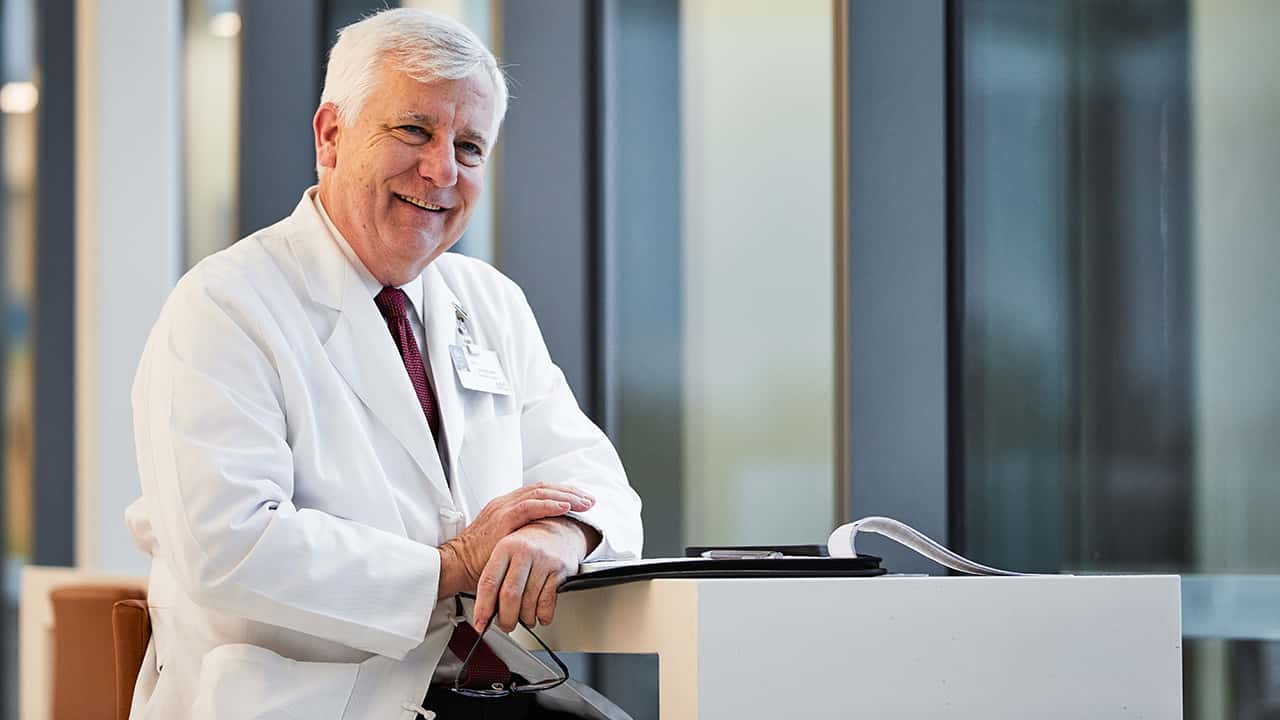 jj_photo_doctor_sitting_to_review_notes_jj_2star Person in weißer Oberbekleidung lehnt an einem Stehpult in einem Innenraum mit großen Fensterflächen. Auf dem Pult liegen Unterlagen; Hintergrund unscharf.
