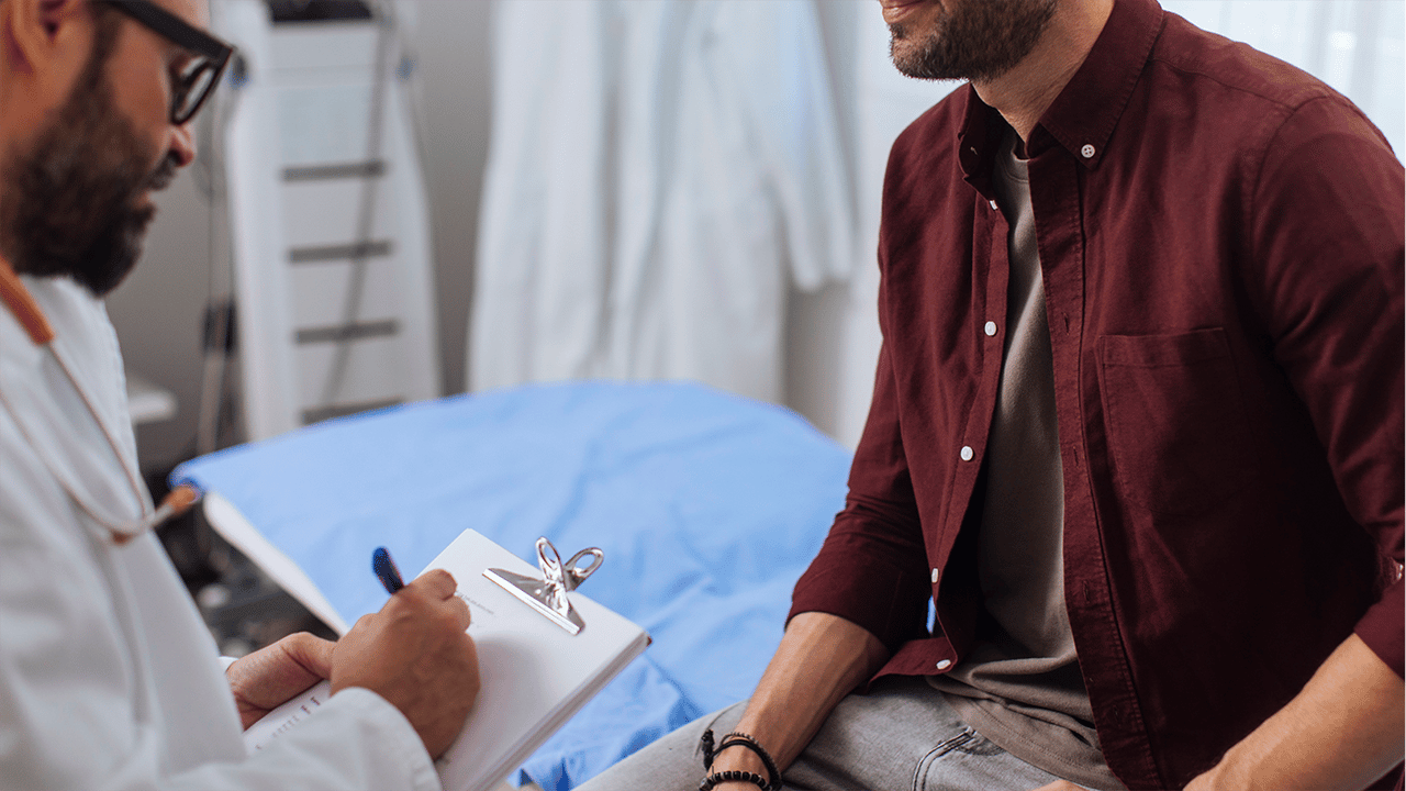 leukamie-und-nachtschweiß-was-kann-ich-tun Zwei Personen befinden sich in einem Innenraum mit Untersuchungsliege. Eine Person schreibt auf ein Klemmbrett, die andere sitzt daneben auf der Liege; medizinische Umgebung sichtbar.