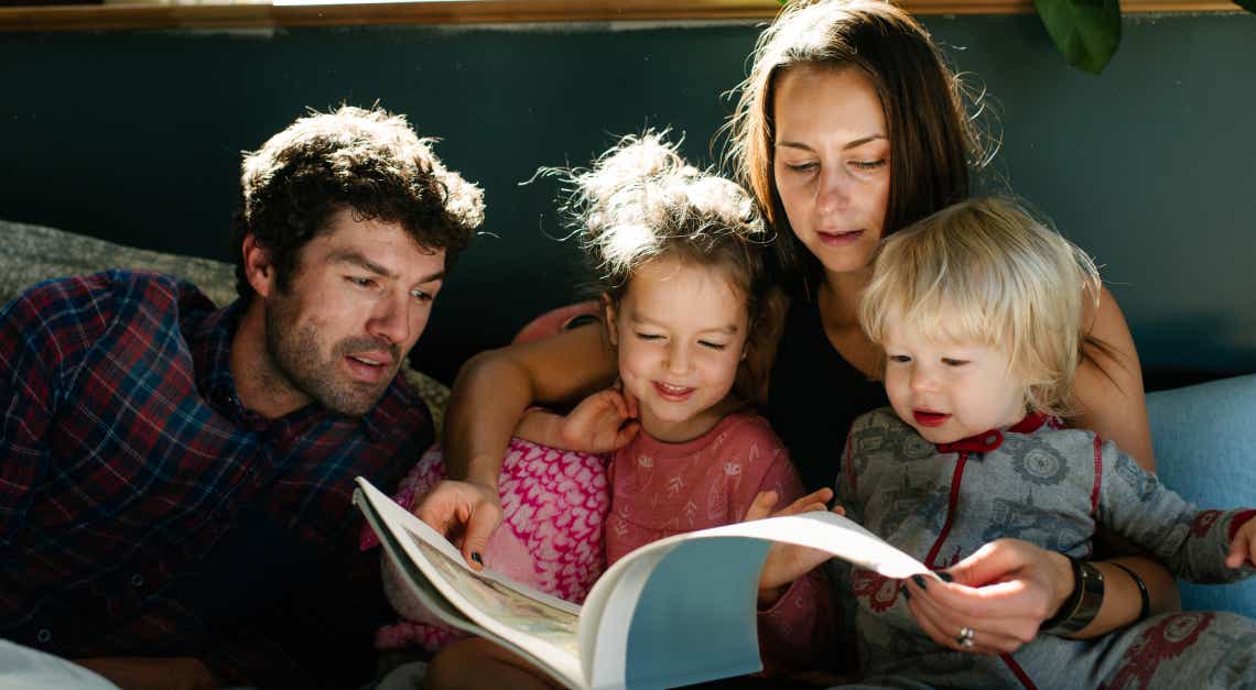 Eine junge vierköpfige Familie sieht sich im Bett bei Tageslicht gemeinsam ein Bilderbuch an.