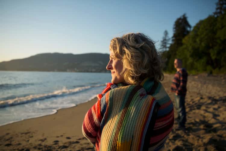 krebserkrankungen-home-header-banner Zwei Personen stehen am Sandstrand nahe der Wasserlinie. Eine Person im Vordergrund ist seitlich zu sehen, eine weitere steht weiter hinten; Meer, Hügel und Bäume im Hintergrund sichtbar.