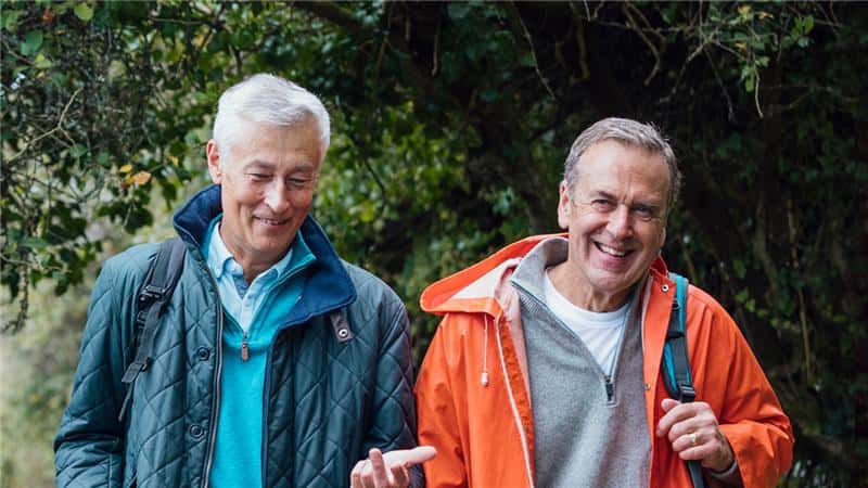 Krebserkrankungen: Blasenkrebs Zwei lächelnde Männer beim Spaziergang im Grünen; einer trägt eine dunkelblaue Steppjacke, der andere eine auffällige orangefarbene Regenjacke.