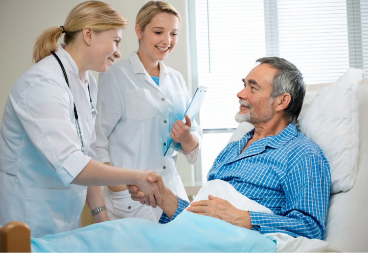 Ärztin und Pflegekraft im Gespräch mit einem älteren Patienten, der in einem Krankenhausbett liegt.  