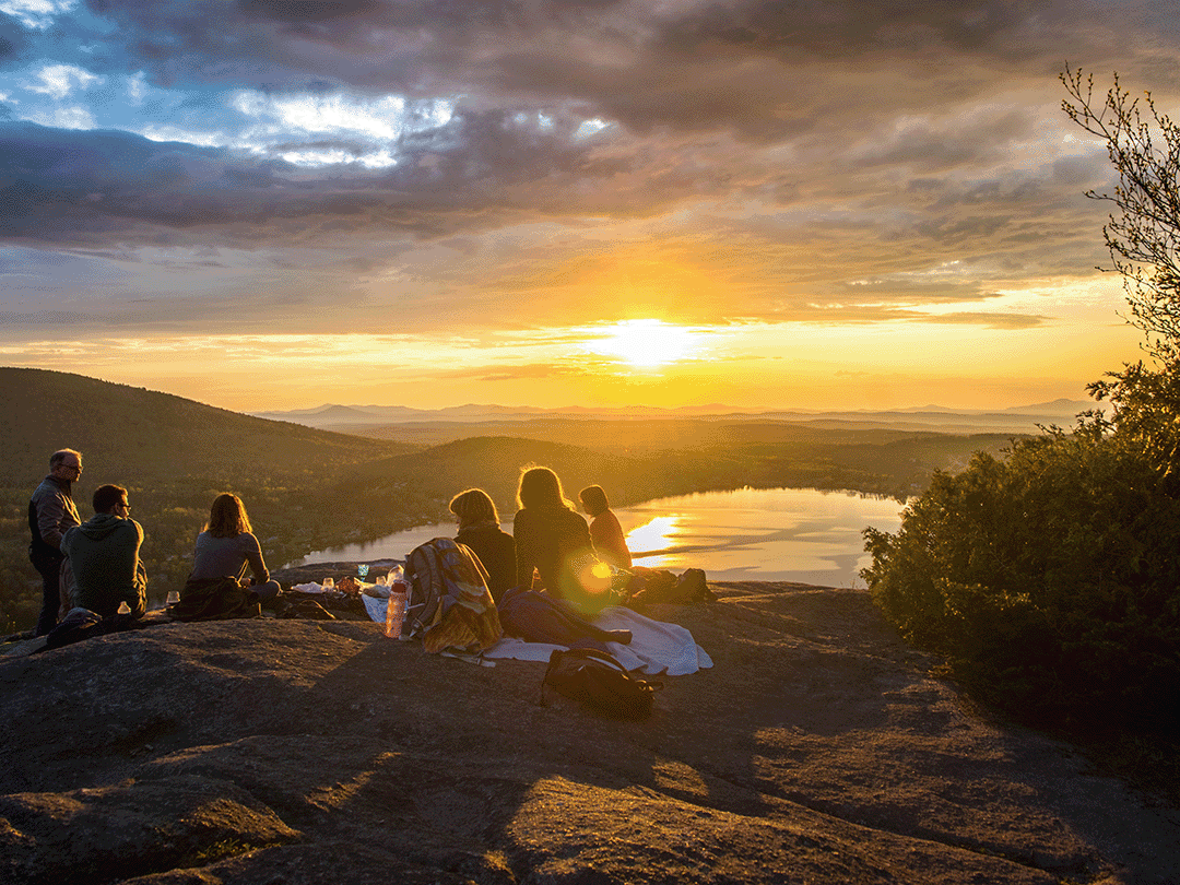 de-de-jwm-ced-mc-hero Freundes-Gruppe mit Morbus Crohn sitzt beim Picknicken auf einem Berg im Sonnenuntergang.