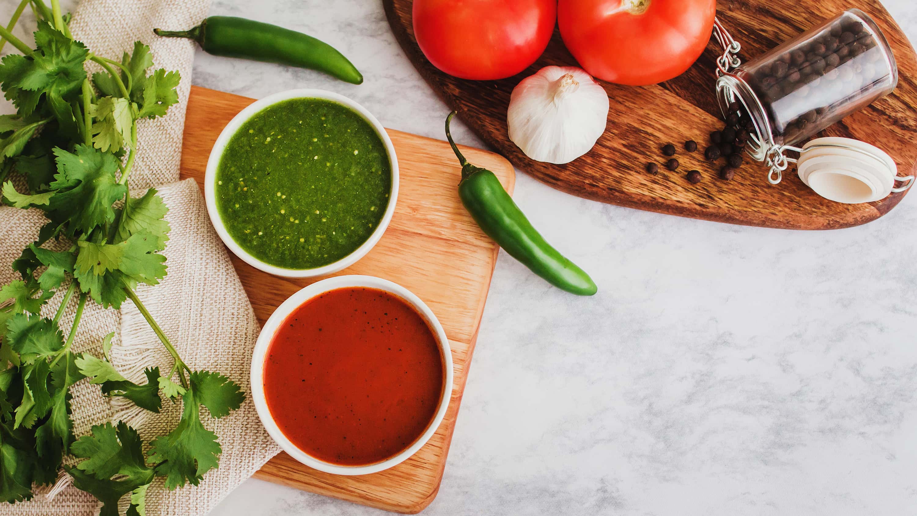 Auf einem Tisch sind Chilischoten, Kräuter, Knoblauch und Tomaten und zwei Schälchen mit roter und grüner Würzsauce. 