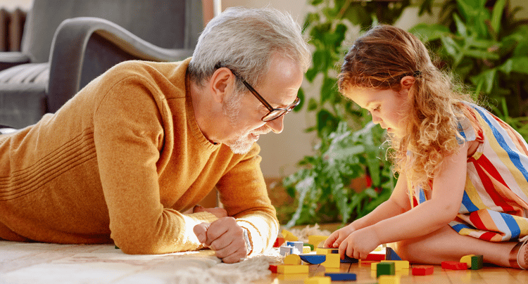 mkr-familie-und-freundeskreis Eine erwachsene Person und ein Kind liegen auf dem Boden und ordnen bunte Bausteine. Innenraum mit Pflanzen, Teppich und Möbeln im Hintergrund sichtbar.