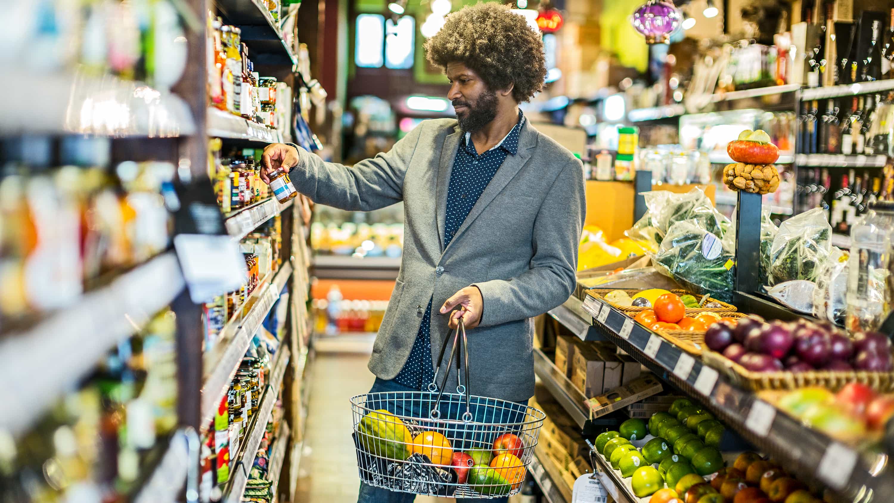 Junger Mann kauft in einem Supermarkt ein. In seinem Korb liegt frisches Obst und Gemüse. Er steht an einem Regal.