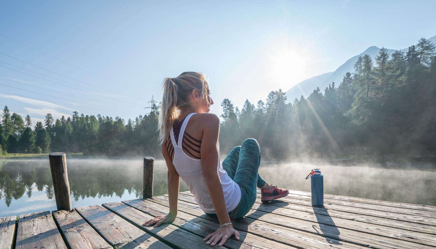 meineced-leben-header_steg Person sitzt seitlich auf einem Holzsteg am See, stützt sich mit den Händen ab. Sportkleidung, Trinkflasche daneben. Im Hintergrund Wasser, Bäume, Berge und tief stehende Sonne.