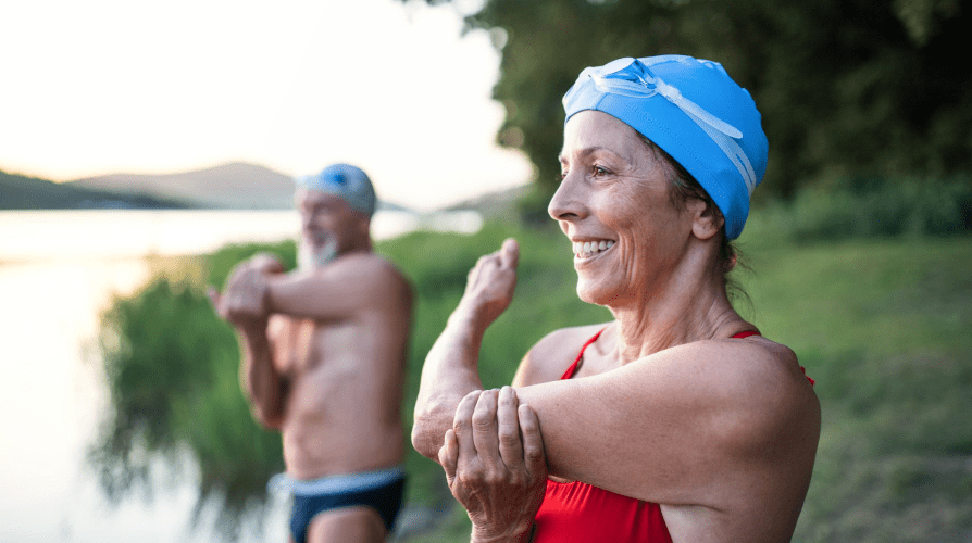 A senior man and woman stretching together near the water during a workout session.