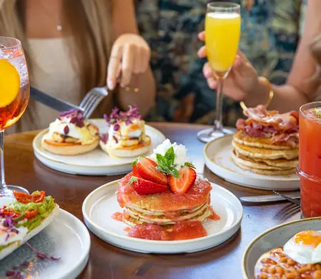 Two friends eating brunch with an assortment of breakfast items on the table, including pancake stacks, and various cocktails