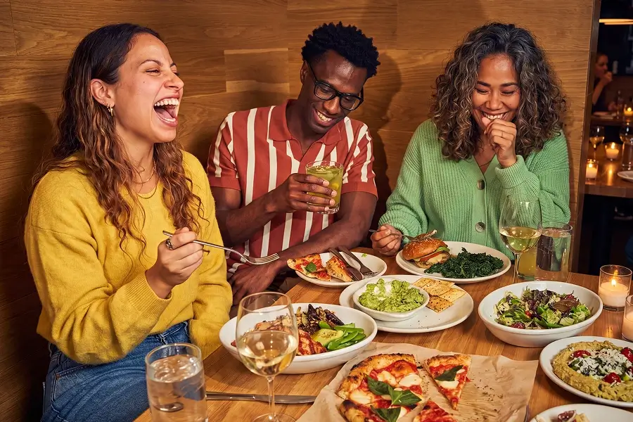 A group of friends out to dinner at Prezzo laughing and enjoying each others company. The ambience is dark and lit with soft fairy lights. Prosecco is also featured on the table alongside the meals.