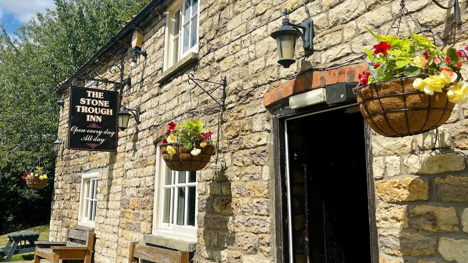 The Stone Trough Inn exterior with stone walls, hanging flower baskets, wooden benches and a traditional pub sign on a sunny day.