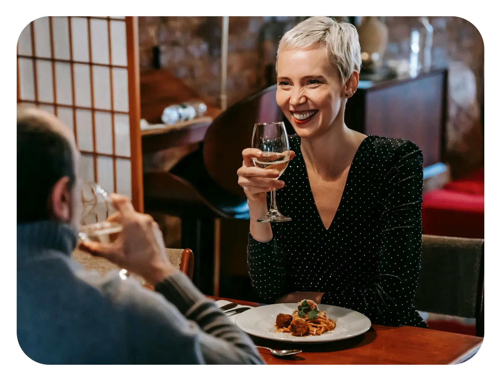 A woman smiling holding a glass of wine with a plate of spaghetti and meatballs in front of her
