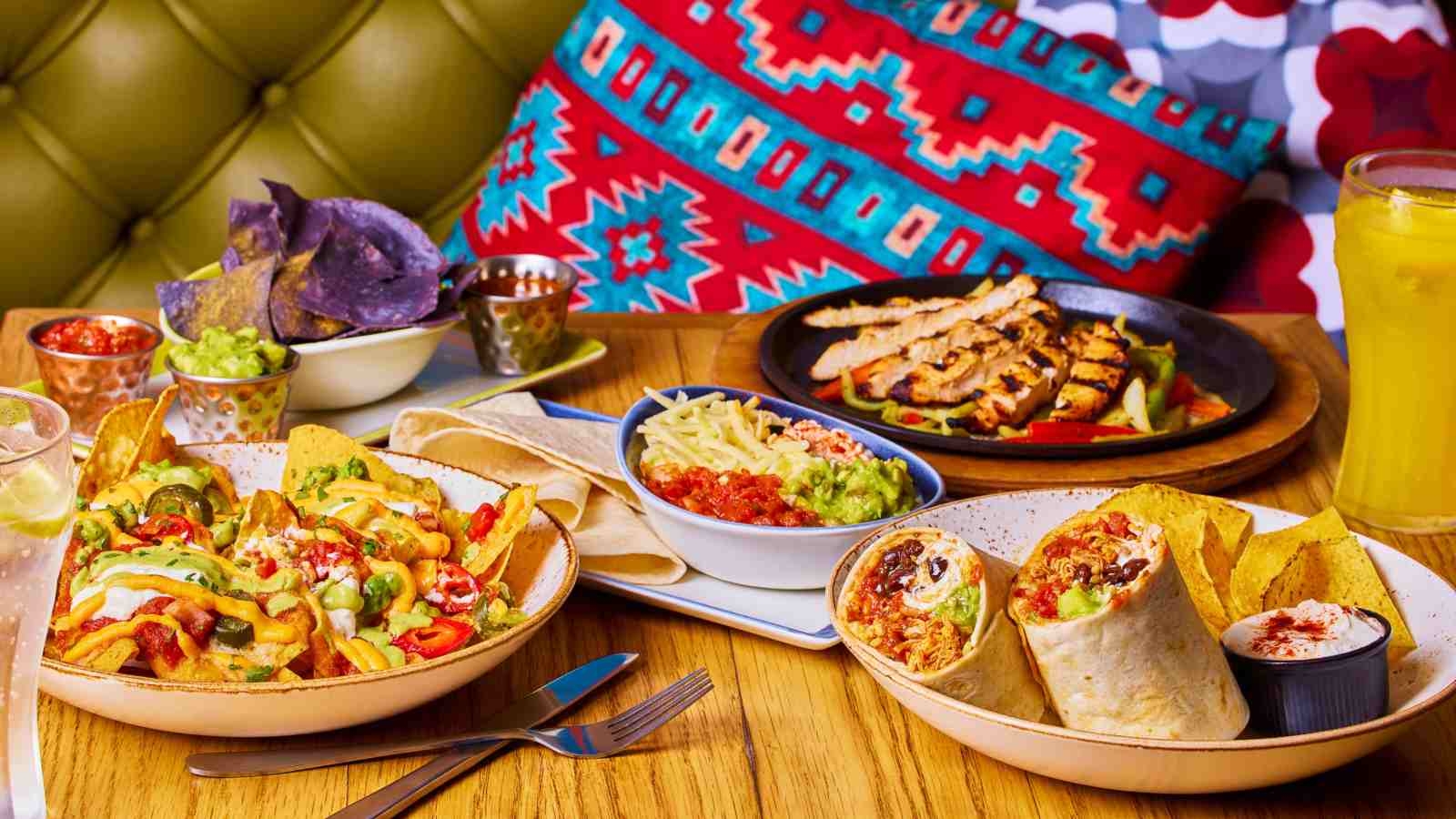 A variety of Mexican food from Chiquito including fries, burritos, tortilla chips and chicken on a table with a patterned cushion in the background