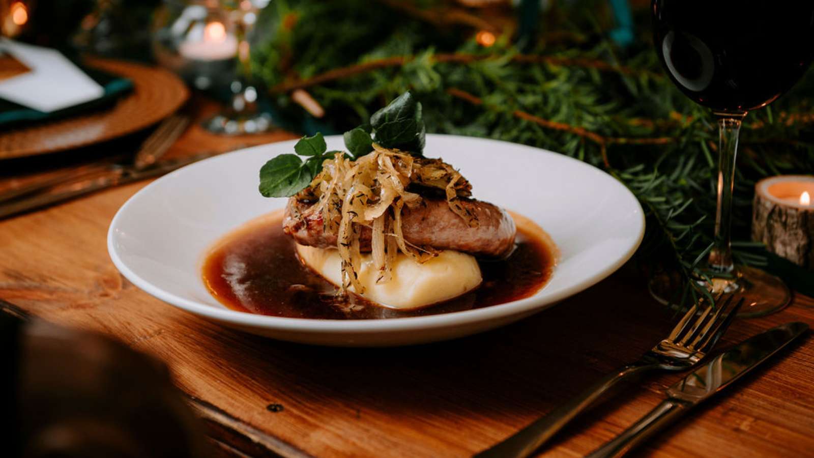 A plate of sausage and mash, topped with onions and onion gravy, set on a wooden table alongside a glass of red wine and an evergreen table centrepiece