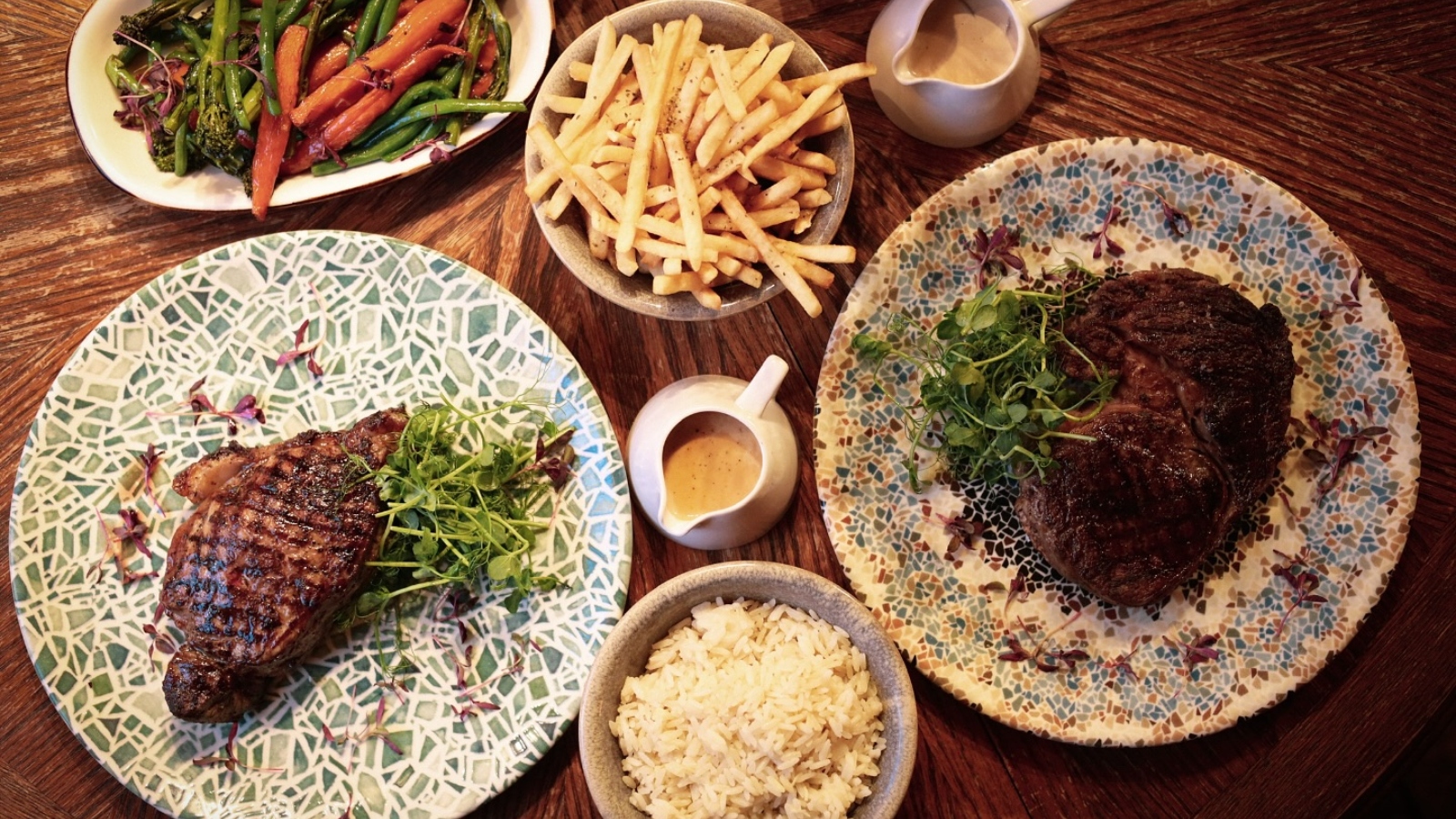Plates of grilled steak served with fries, rice, vegetables and sauces on a wooden table.