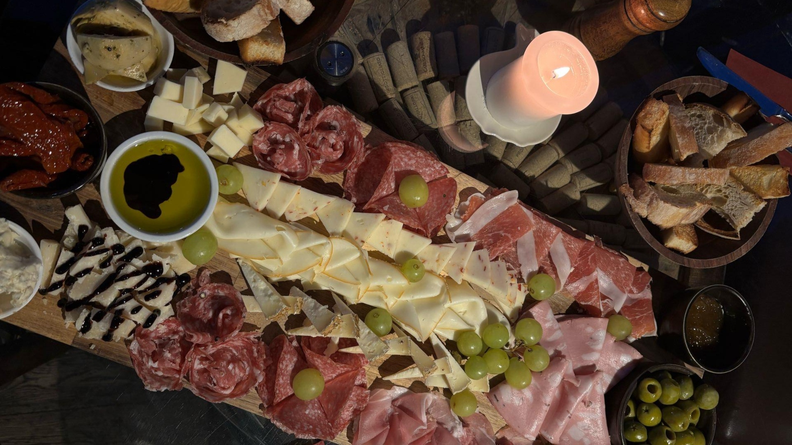 A large charcuterie board with sliced meats, cheeses, grapes, olives, bread, dips, and a lit candle in the center.