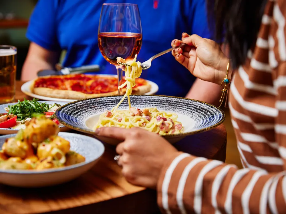 Close-up of a woman twirling spaghetti carbonara around her fork while dining at a restaurant, with a glass of rosé wine and a pizza blurred in the background.