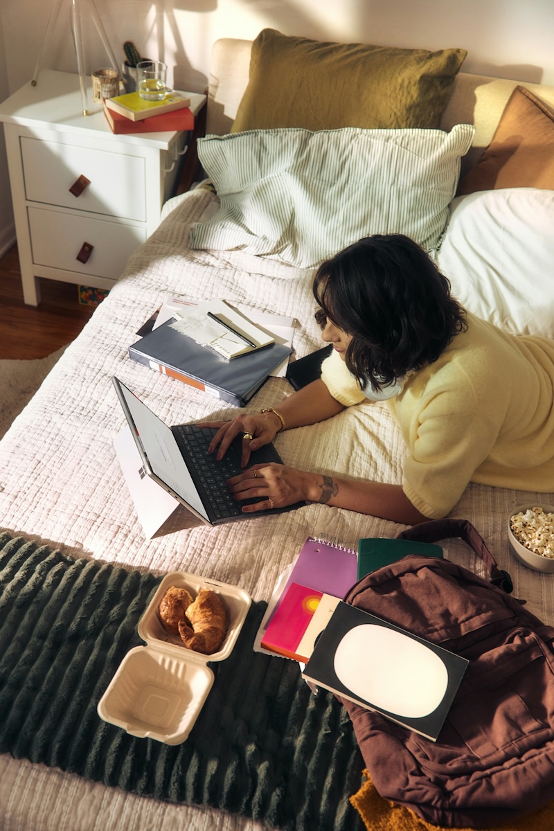 Student laid on the bed typing on her laptop surrounded by notebooks, files, her backpack and snacks