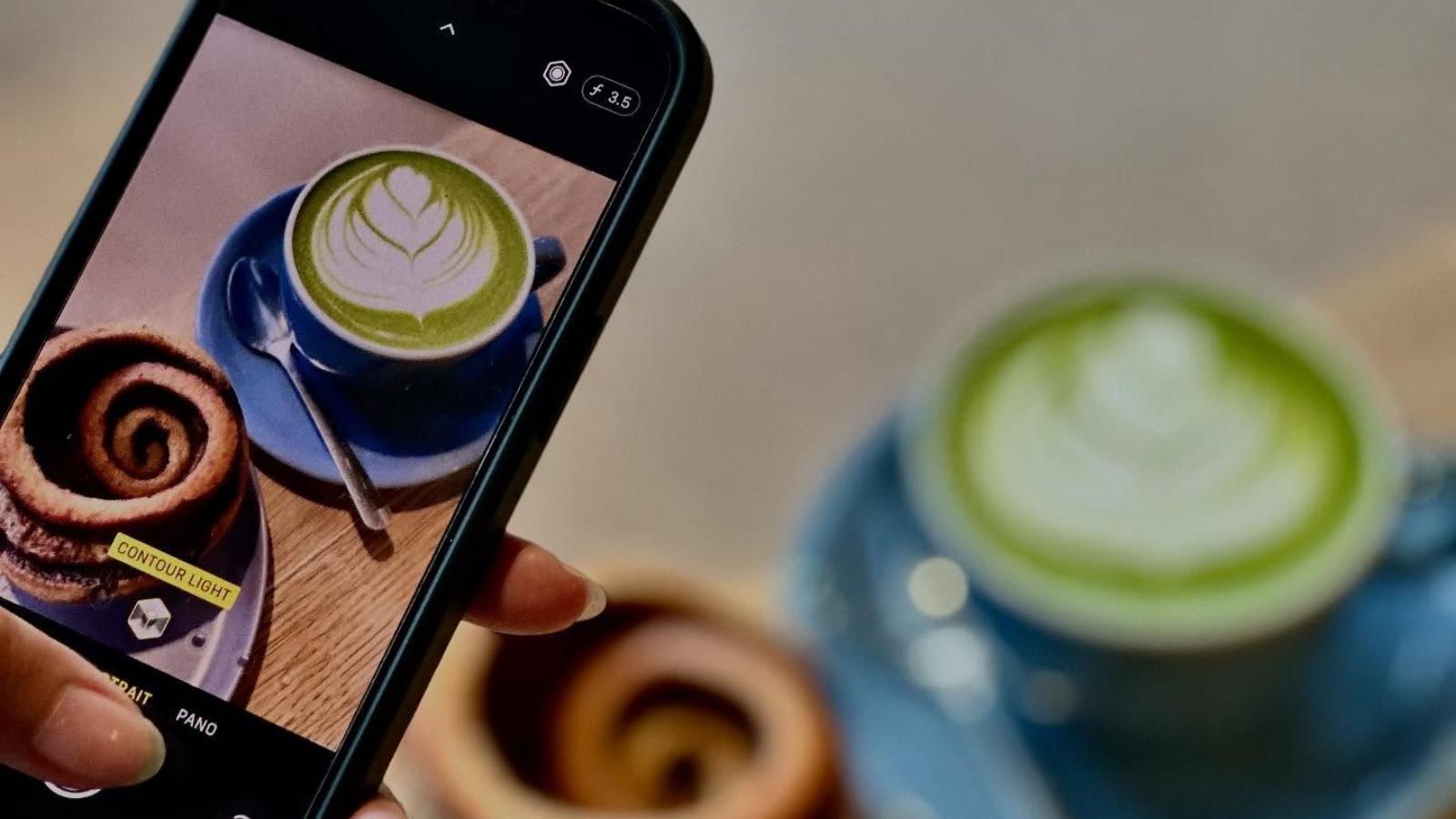 A phone taking a photo of a matcha latte and a pastry, with the real matcha latte and pastry blurred in the background.