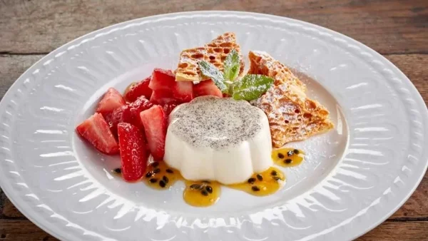 A panna cotta presented neatly on a white ceramic plate, topped with strawberries, passionfruit juices, and mint. 