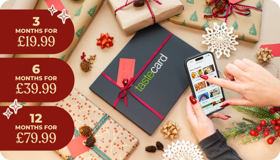 “A tastecard gift box with a bow surrounded by Christmas presents, while someone nearby uses the tastecard app on their phone.