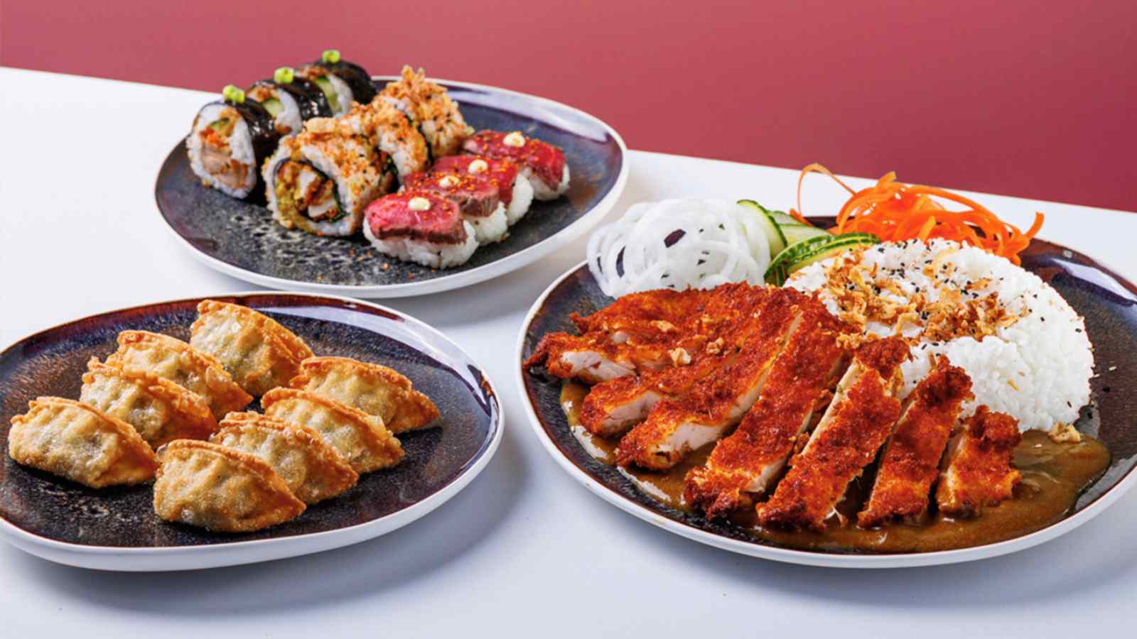 Three plates of food on a white table. One of chicken katsu curry served with sushi rice, one of gyoza and one of sushi, featuring crispy duck futomaki, spicy chicken katsu iso rolls, and seared beef nigiri