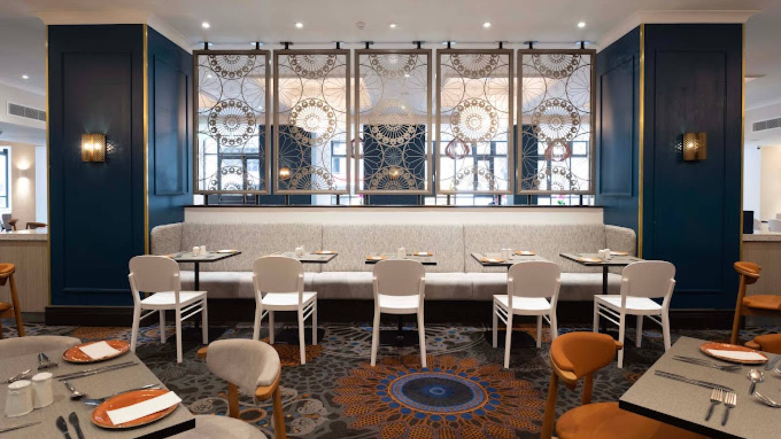 Modern restaurant interior with patterned room divider, wall panels, white chairs, and neatly set tables, all in tones of white, blue, and orange.