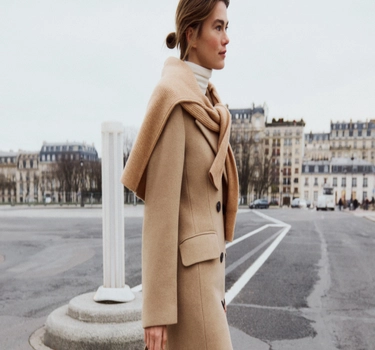 A young woman walking in a city wearing a white sweater, and long brown sleeve jacket for the Fall season from Companys. 