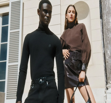 A young man wearing black jeans and black trouser in front of a woman. A young woman with brown hair wearing a brown sweater and black leathered skirt. 