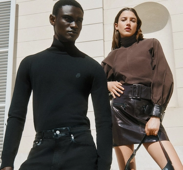 A young man wearing black jeans and black trouser in front of a woman. A young woman with brown hair wearing a brown sweater and black leathered skirt.