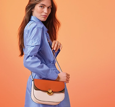 A young woman with long brown hair. Woman wearing a blue long-sleeve dress and wearing a beige and red bag from Furla.